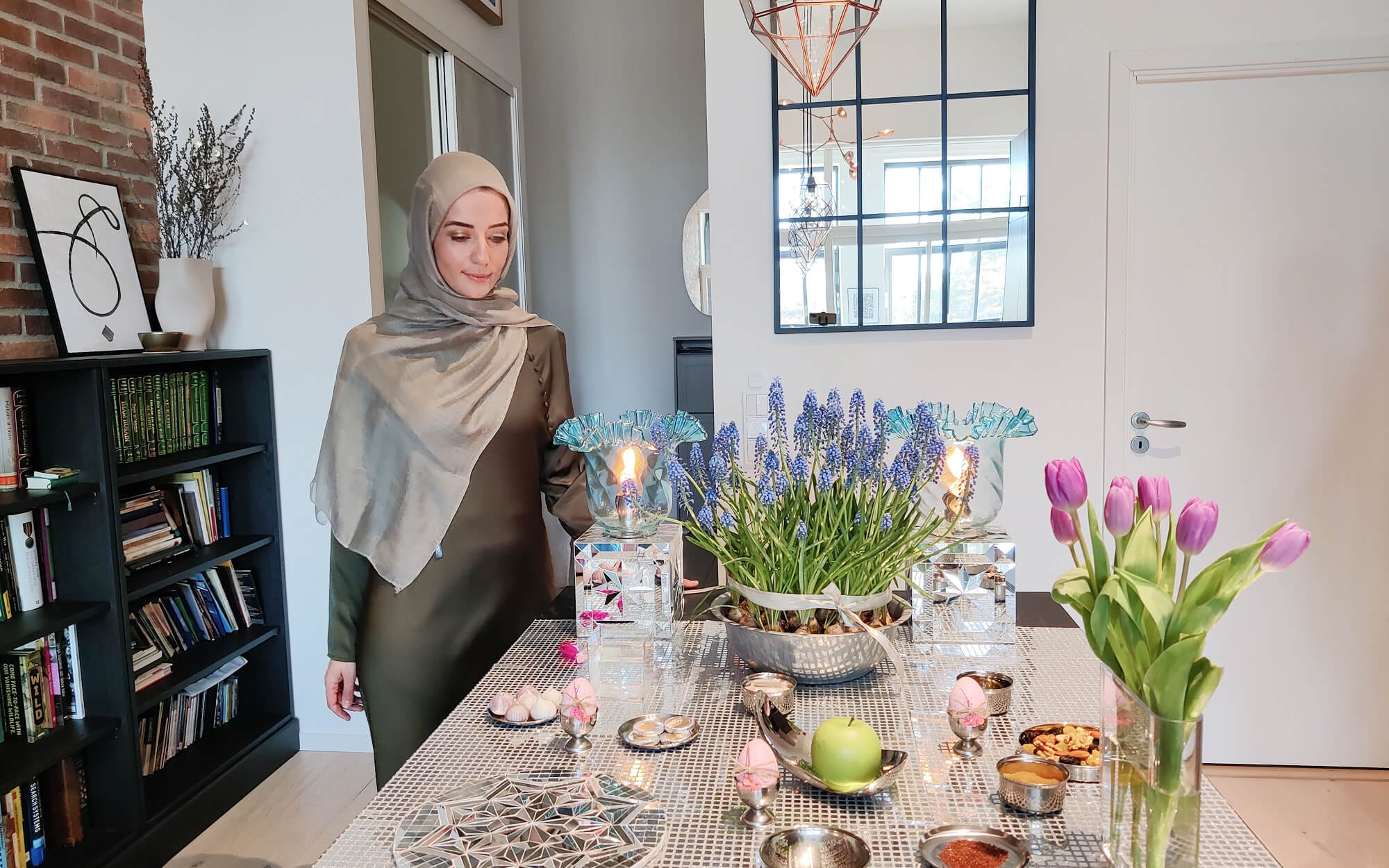 A Haftseen table arranged on a reflective mirror-mosaic surface, with painted eggs, candles, fresh greens, fruit and flowers carefully placed in small metal dishes.