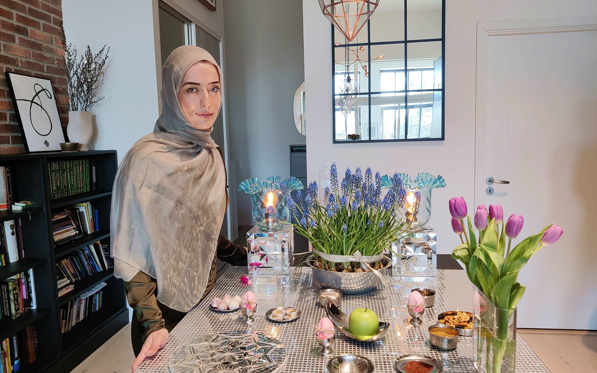 A Haftseen table arranged on a reflective mirror-mosaic surface, with painted eggs, candles, fresh greens, fruit and flowers carefully placed in small metal dishes.