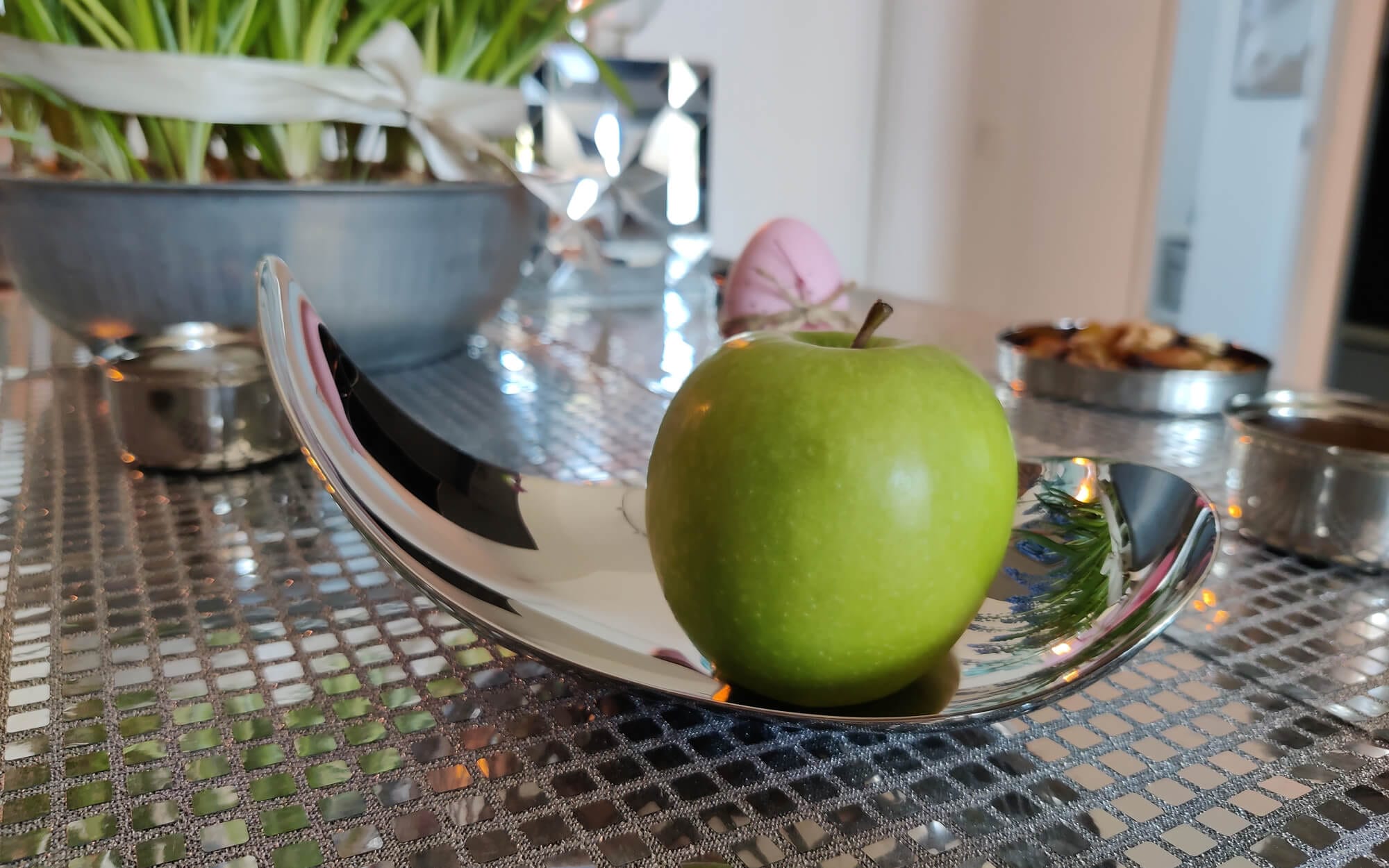 A single green apple resting on a curved, polished metal dish on a mirrored Haftseen table.