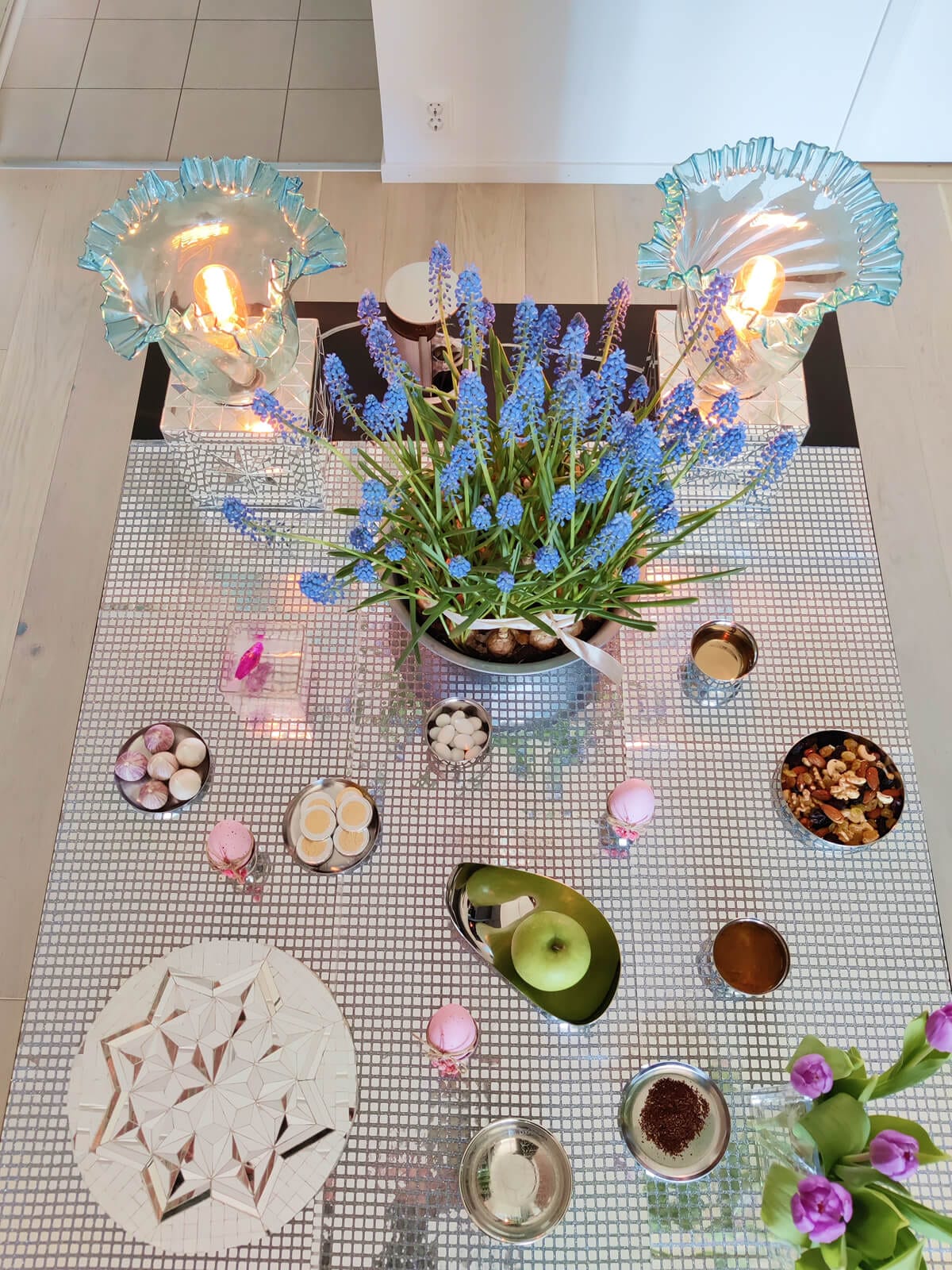 A mirrorwork-inspired Haftseen table, arranged on a reflective mosaic surface with blue hyacinths, painted eggs, a green apple, nuts, and glowing glass candle holders.