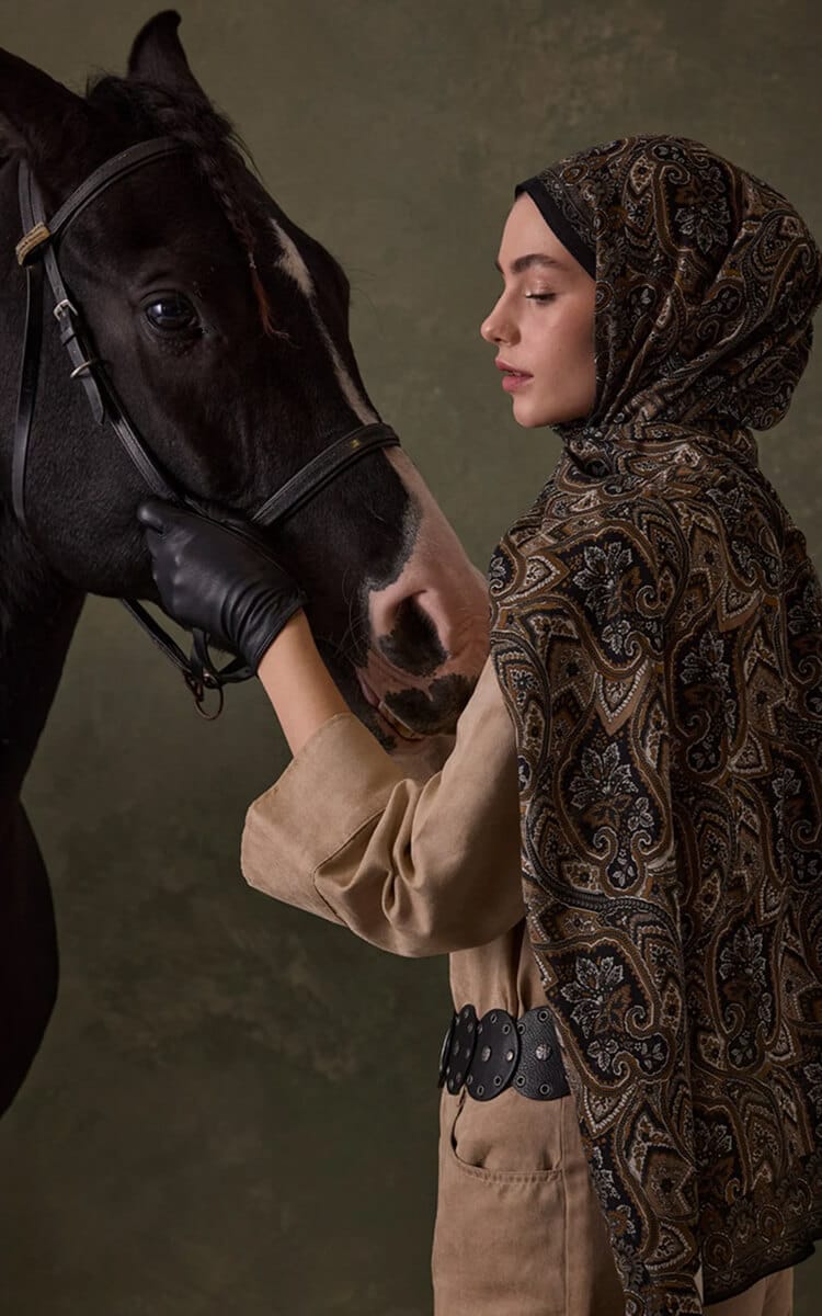 Woman in a paisley hijab gently holding a black horse’s face.