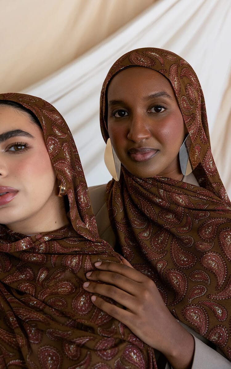 Two women wearing matching brown paisley-patterned hijabs, standing close together in front of a softly draped light fabric backdrop.