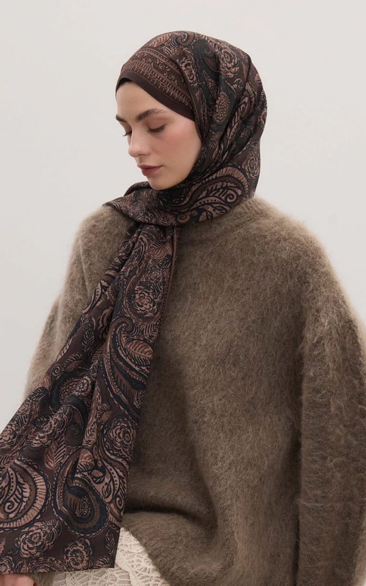 Woman wearing a dark paisley-patterned hijab and a soft brown sweater, looking down in profile.