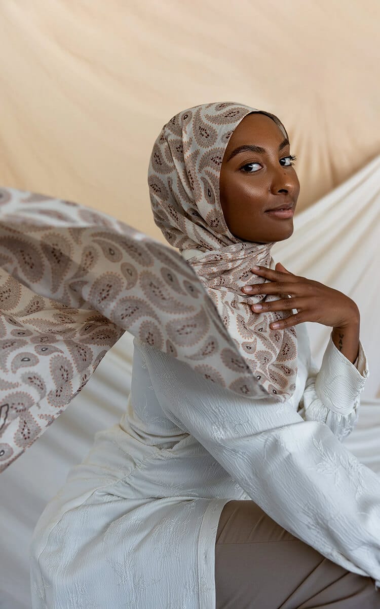A woman wearing a patterned beige paisley hijab and a light-coloured dress, seated with the fabric of her scarf flowing to the side.