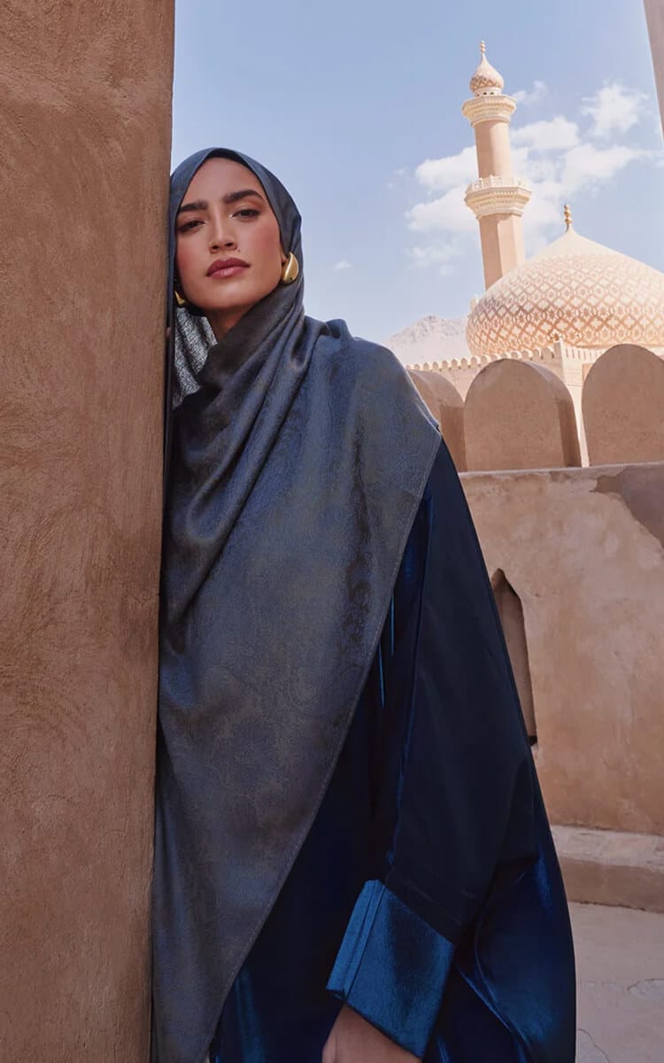 Woman wearing a blue pashmina-style hijab, standing beside a mosque.