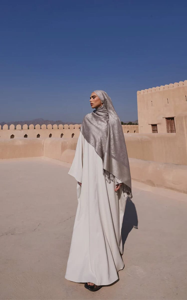 Woman wearing a silver-grey pashmina over a long white dress, standing in a desert courtyard with traditional mud-brick architecture.