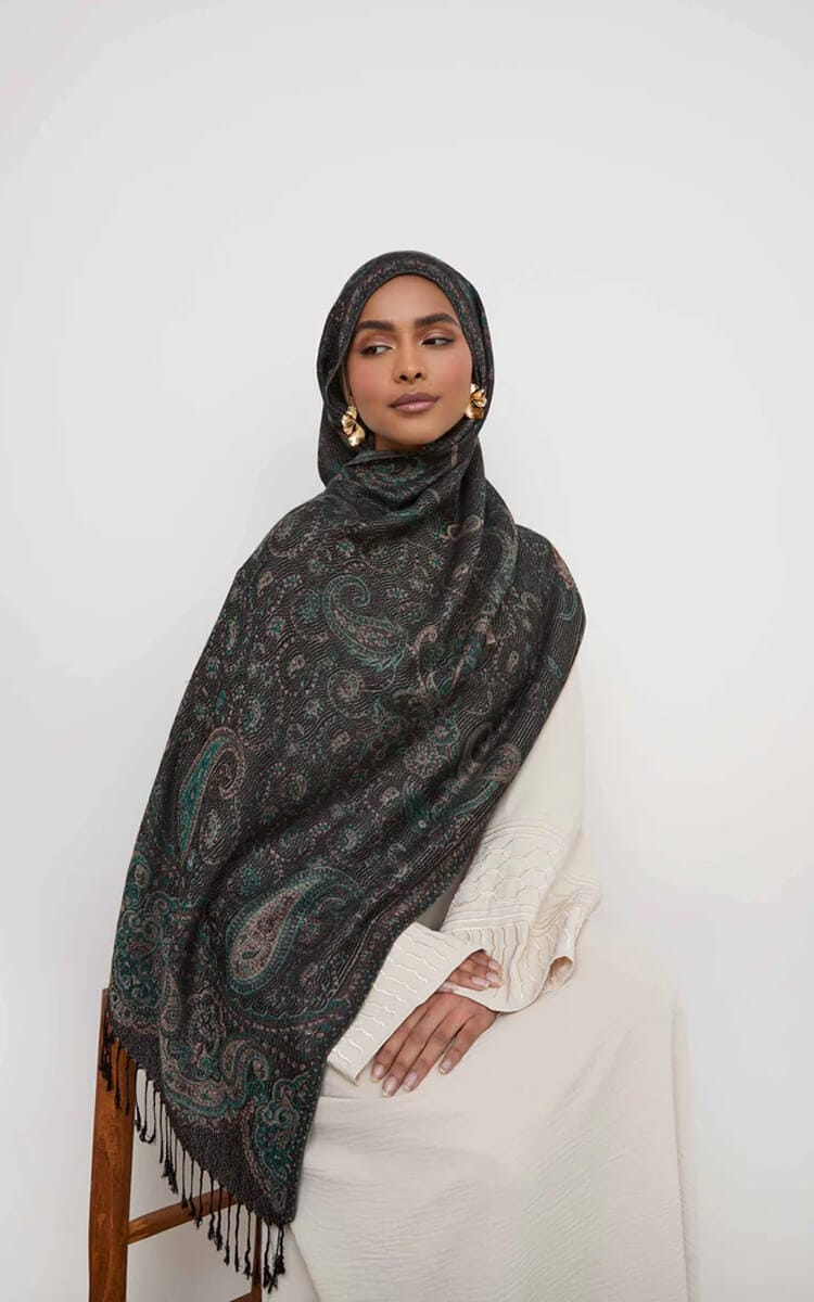 A woman wearing a dark pashmina with an intricate paisley pattern, draped as a hijab, sitting gracefully on a wooden chair against a plain light background.