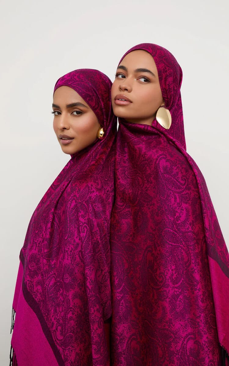 Two women wearing rich magenta pashmina scarves with an intricate paisley pattern, styled as hijabs