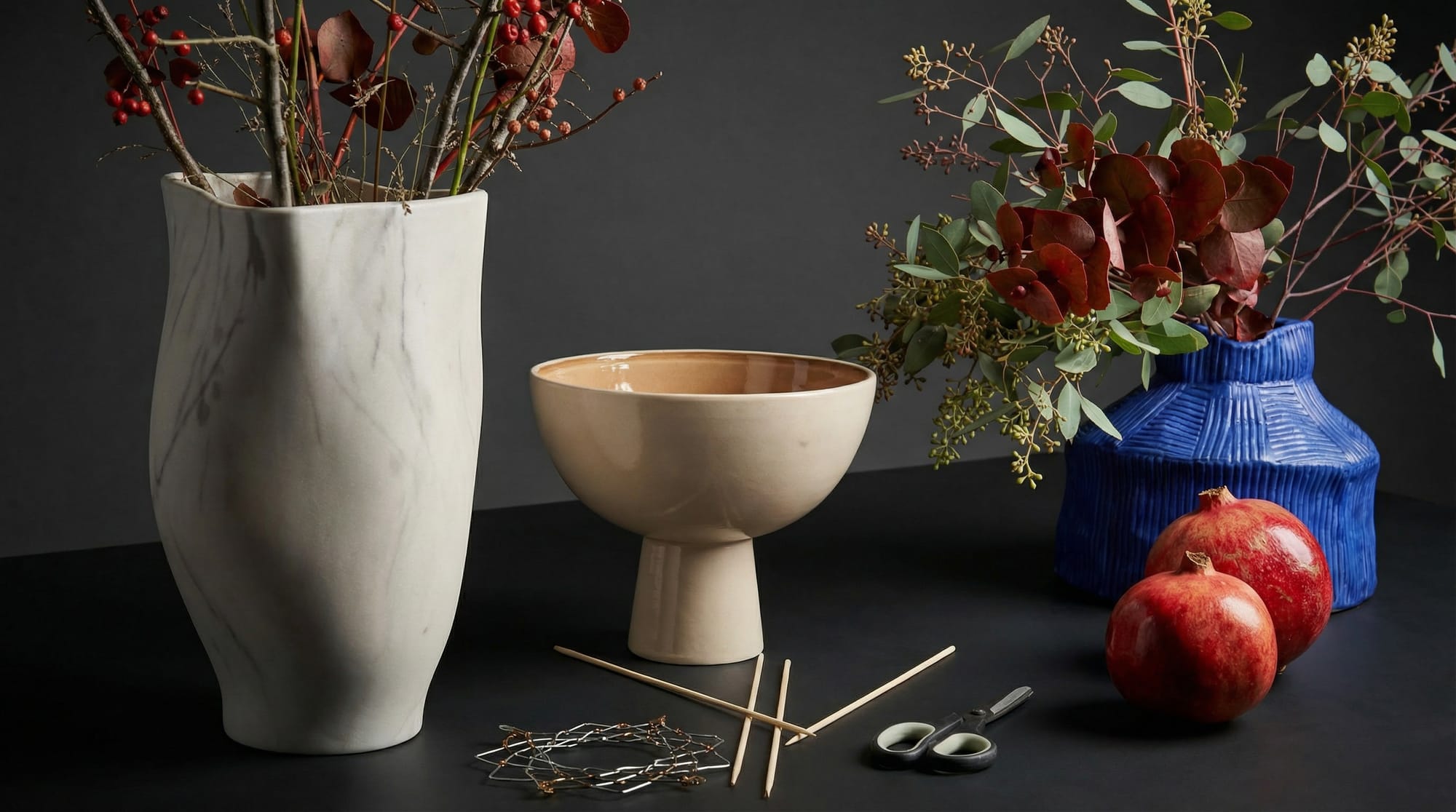 A still life of vases, greenery, red berries, and pomegranates laid out with floral tools for creating a winter pomegranate arrangement.