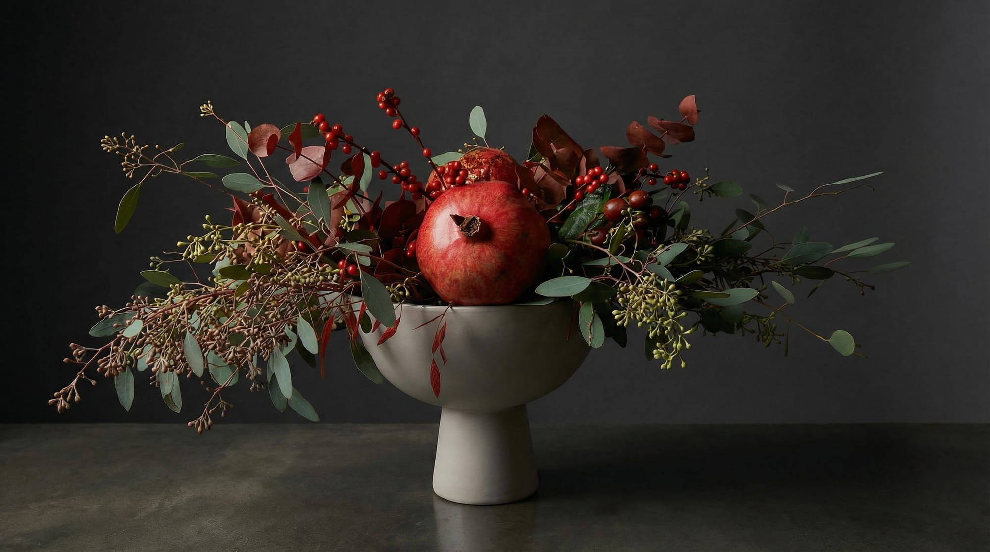 Pomegranate centerpiece with seasonal branches and greenery, styled for Yalda Night on a minimal pedestal bowl.