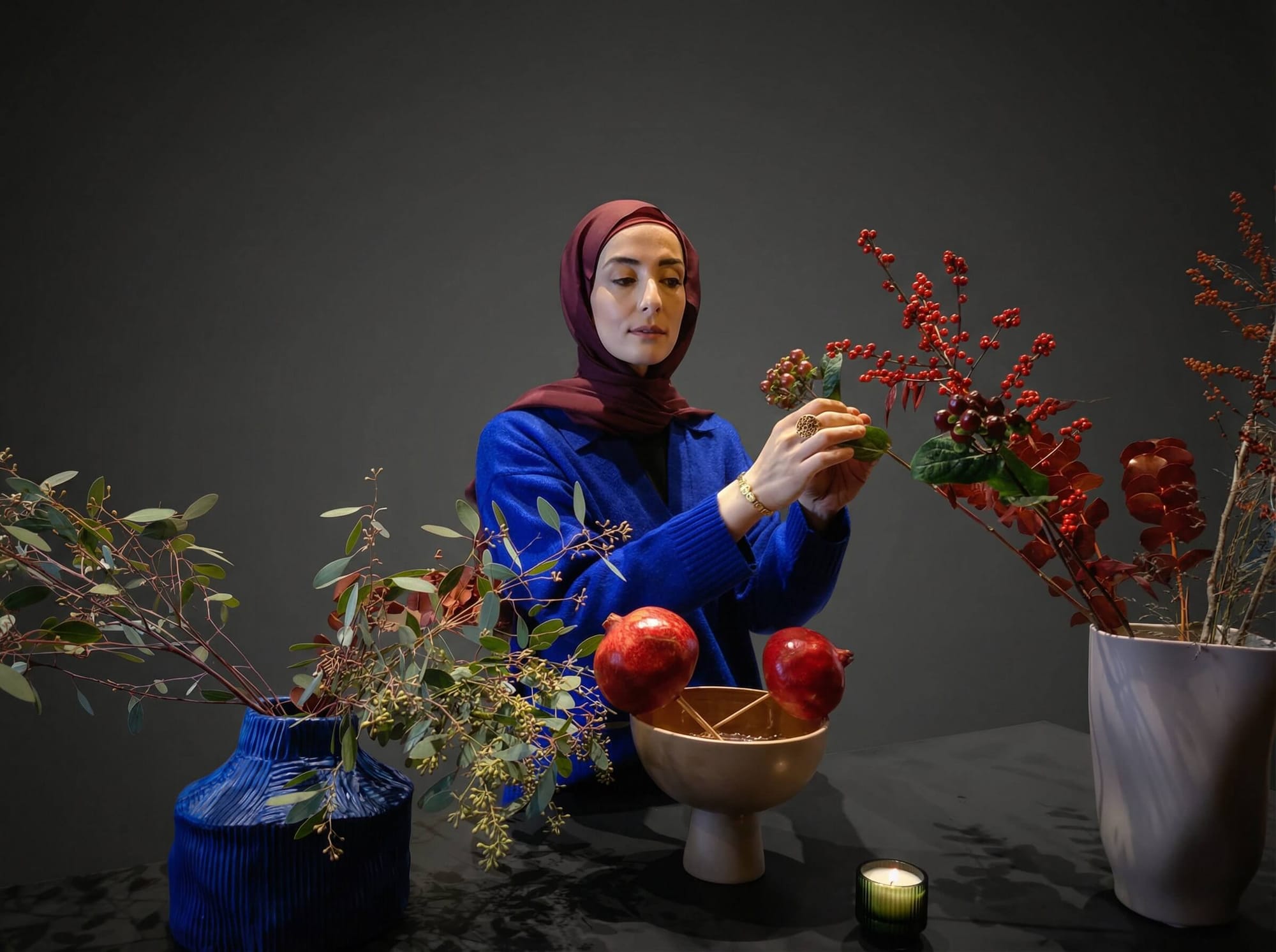 A woman in a burgundy hijab arranging a pomegranate and winter foliage centerpiece with red berries and eucalyptus.