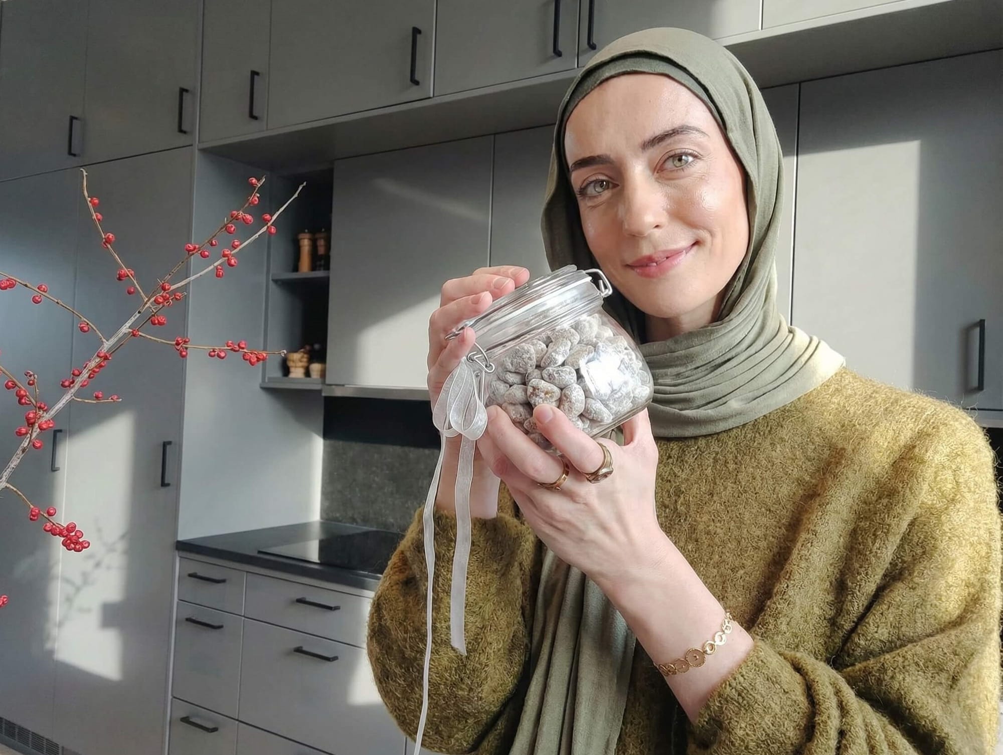 Person wearing an olive hijab holding a glass jar filled with coated almonds in a modern grey kitchen.