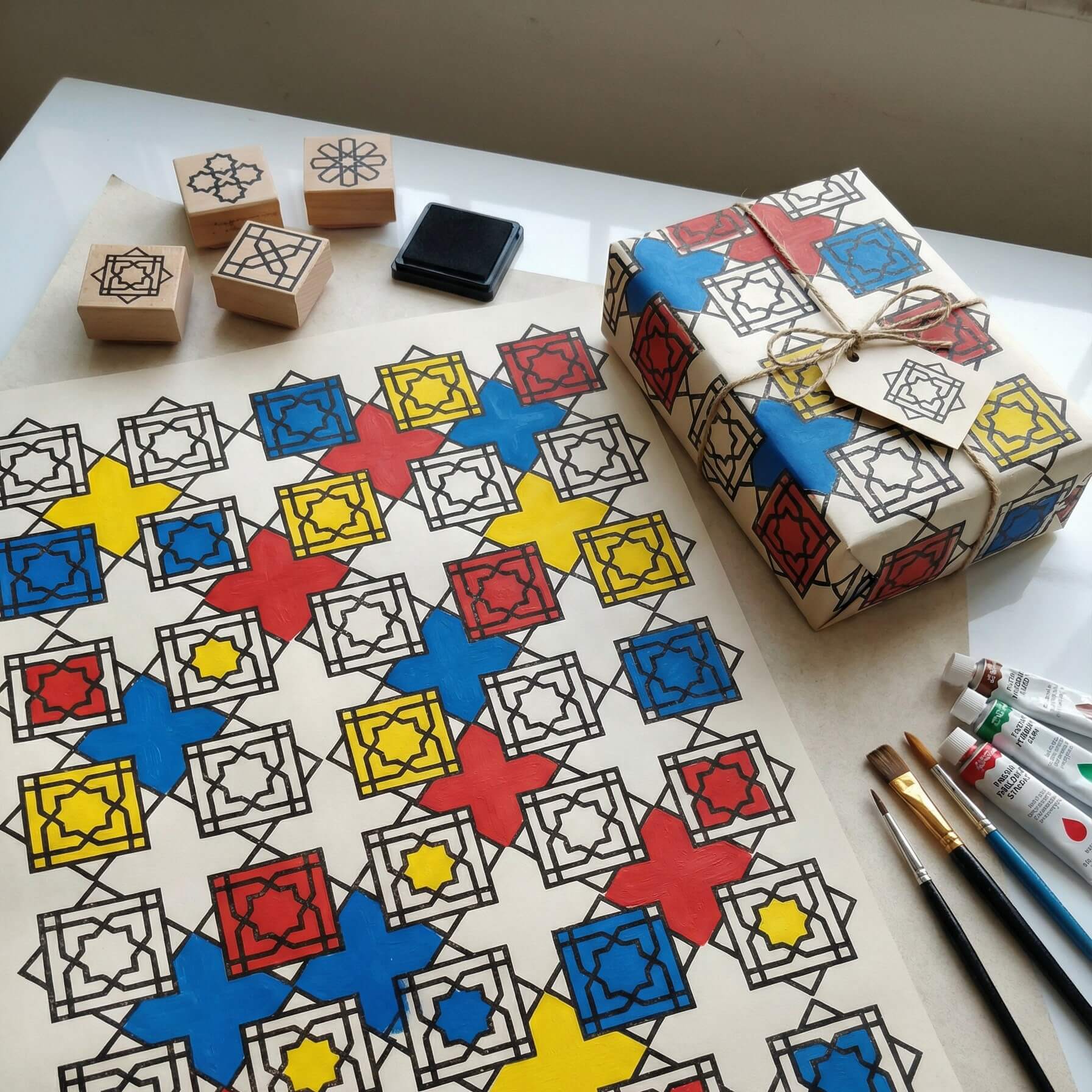 DIY Mondrian-inspired geometric gift wrap created with Islamic pattern stamps, hand-painted in red, blue, and yellow using acrylic paint, shown with wrapped gift and art tools.
