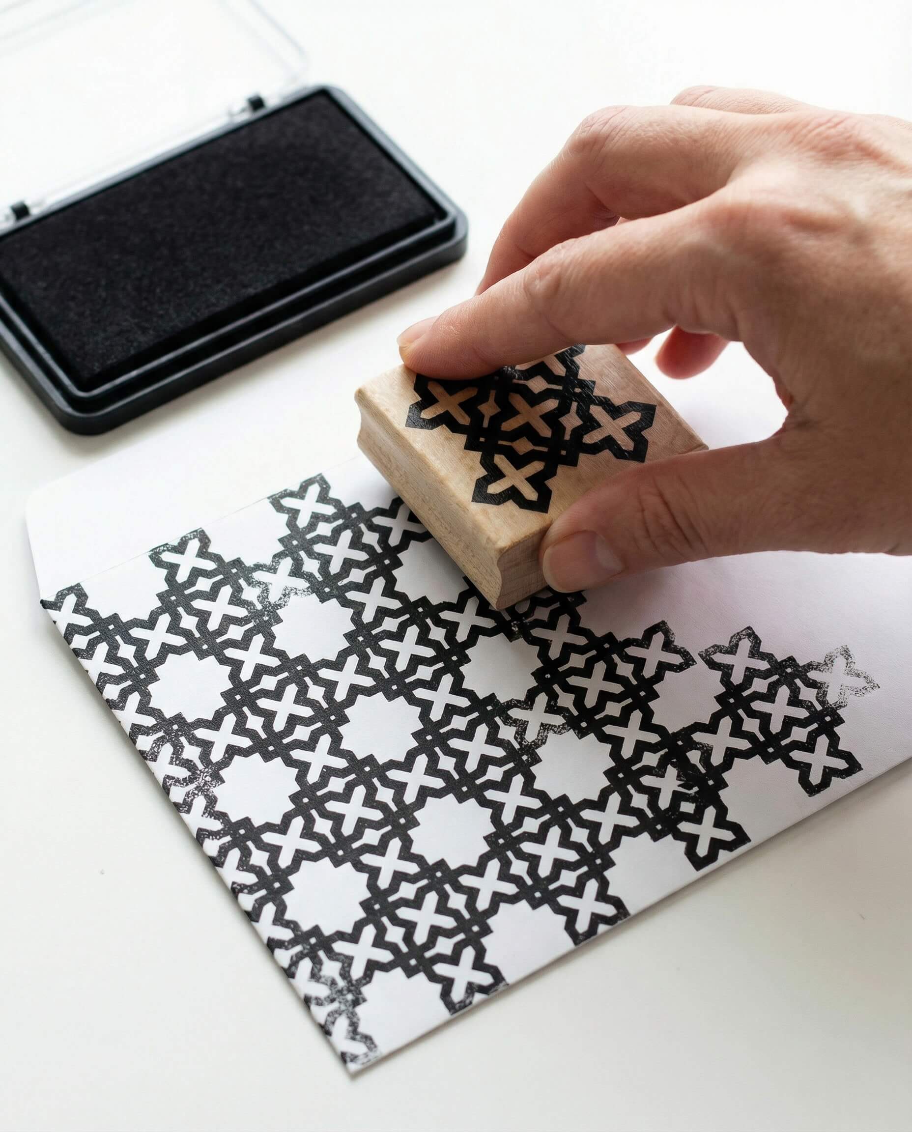 Stamping an Islamic geometric pattern onto a paper envelope using a wooden stamp and black ink pad.