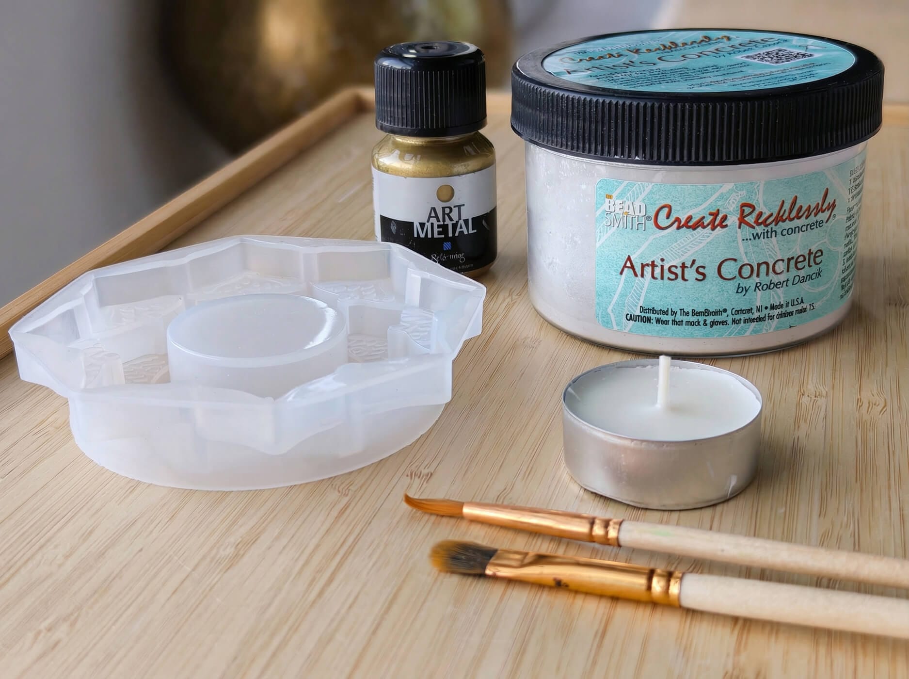 Materials for making a concrete candleholder DIY, including decorative concrete powder, silicone mold, gold paint, and tealight candle