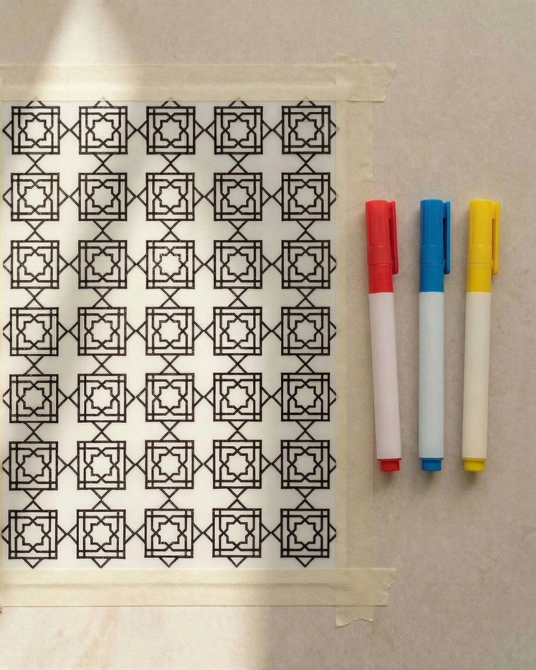 Stamped Islamic geometric pattern repeated in a structured grid on acrylic paper, taped edges prepared for colouring with red, blue, and yellow acrylic markers.
