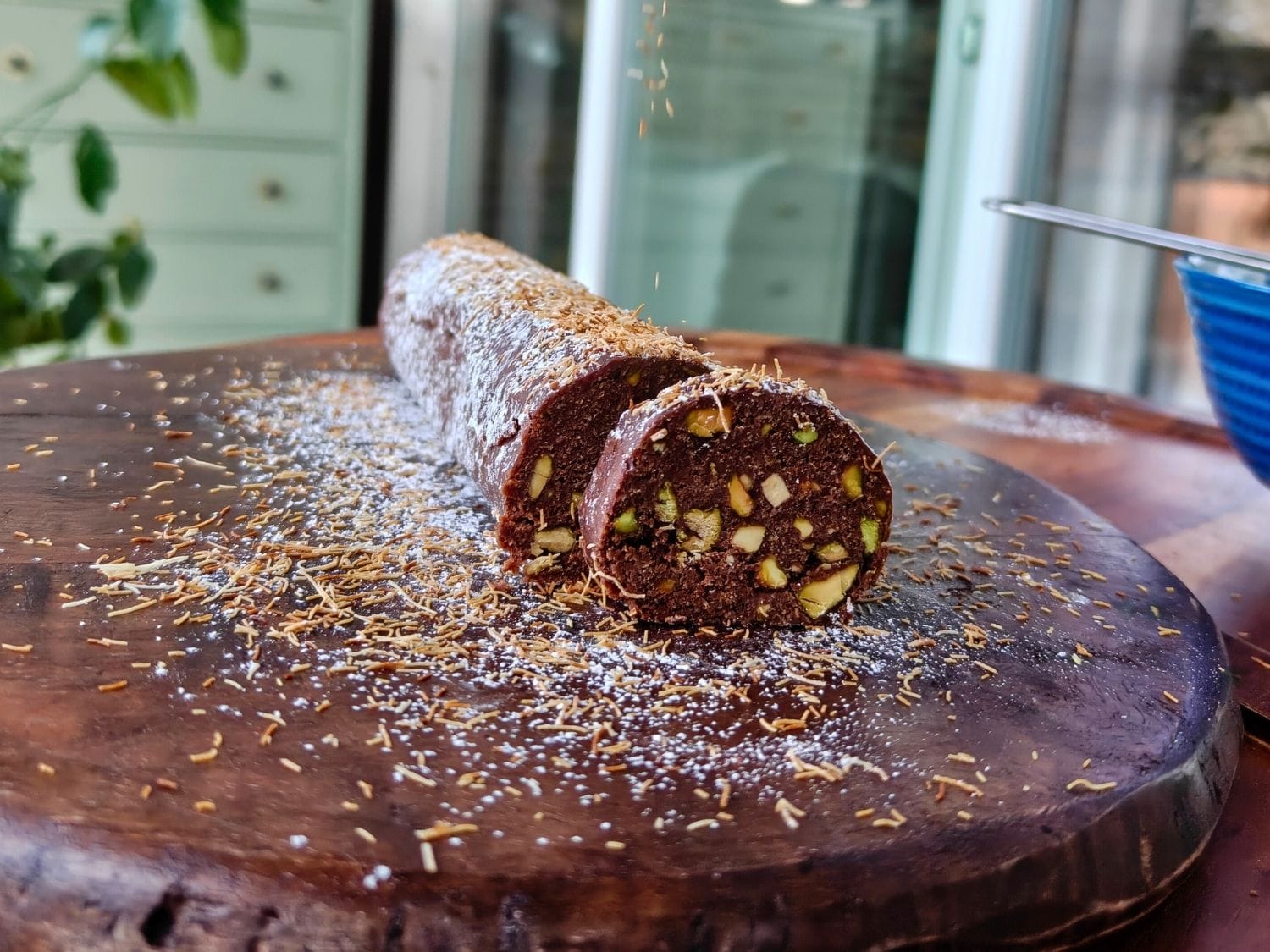 Sliced chocolate kataifi salami with pistachios, dusted with powdered sugar and toasted kataifi on a wooden board.