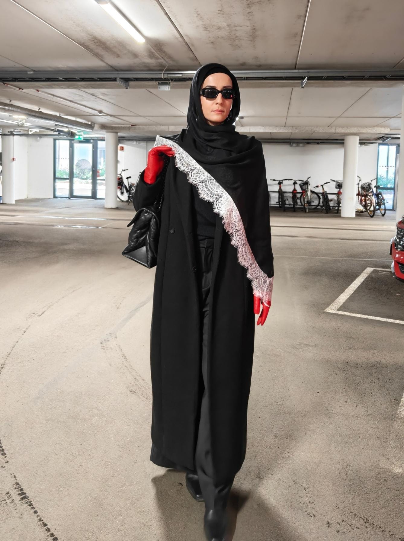 Woman wearing a black modal hijab with off-white lace derail with a long black coat and red gloves in a modern modest outfit