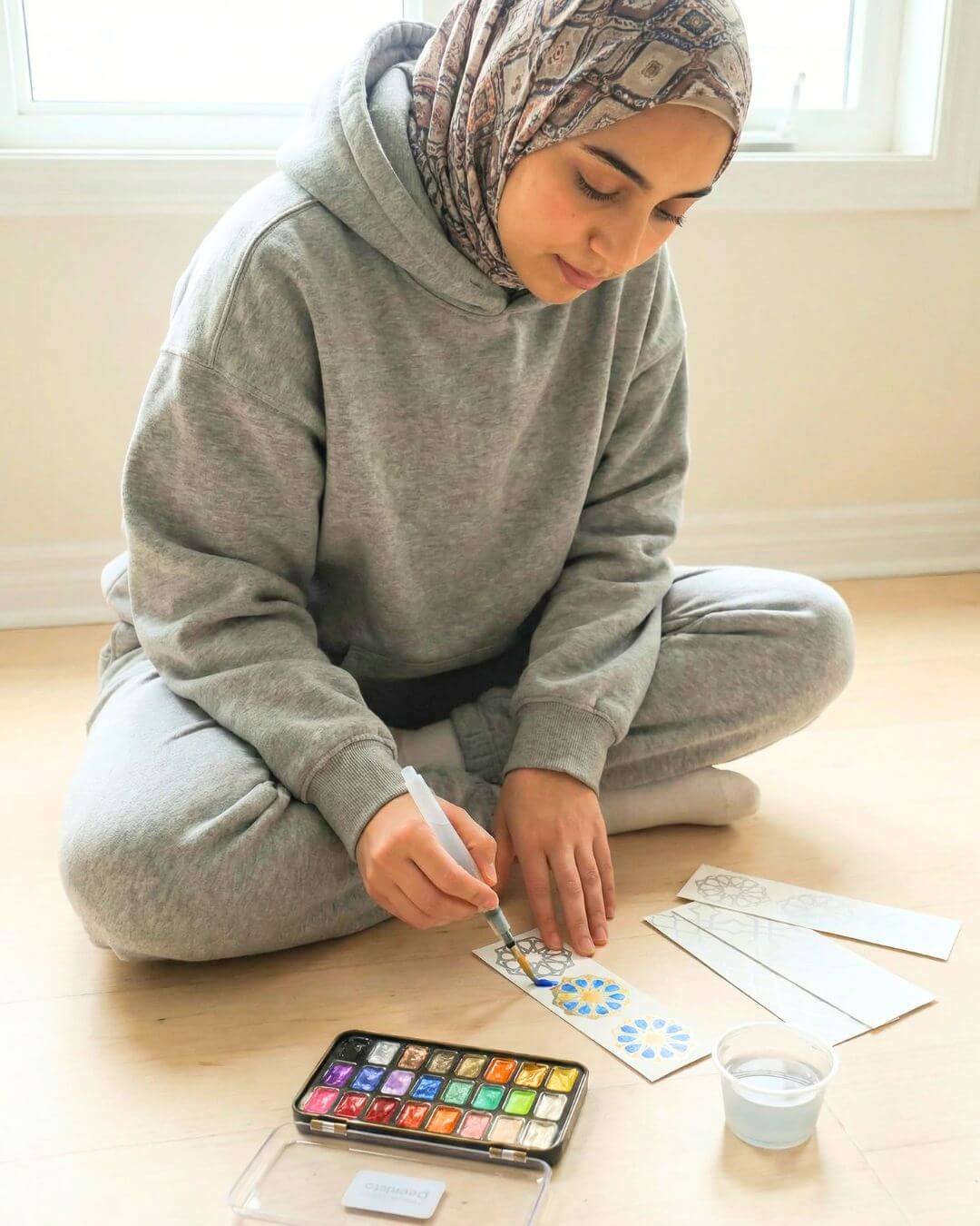 Muslim teen girl painting an Islamic bookmark with the Deenista shimmer watercolor kit while sitting on the floor at home.