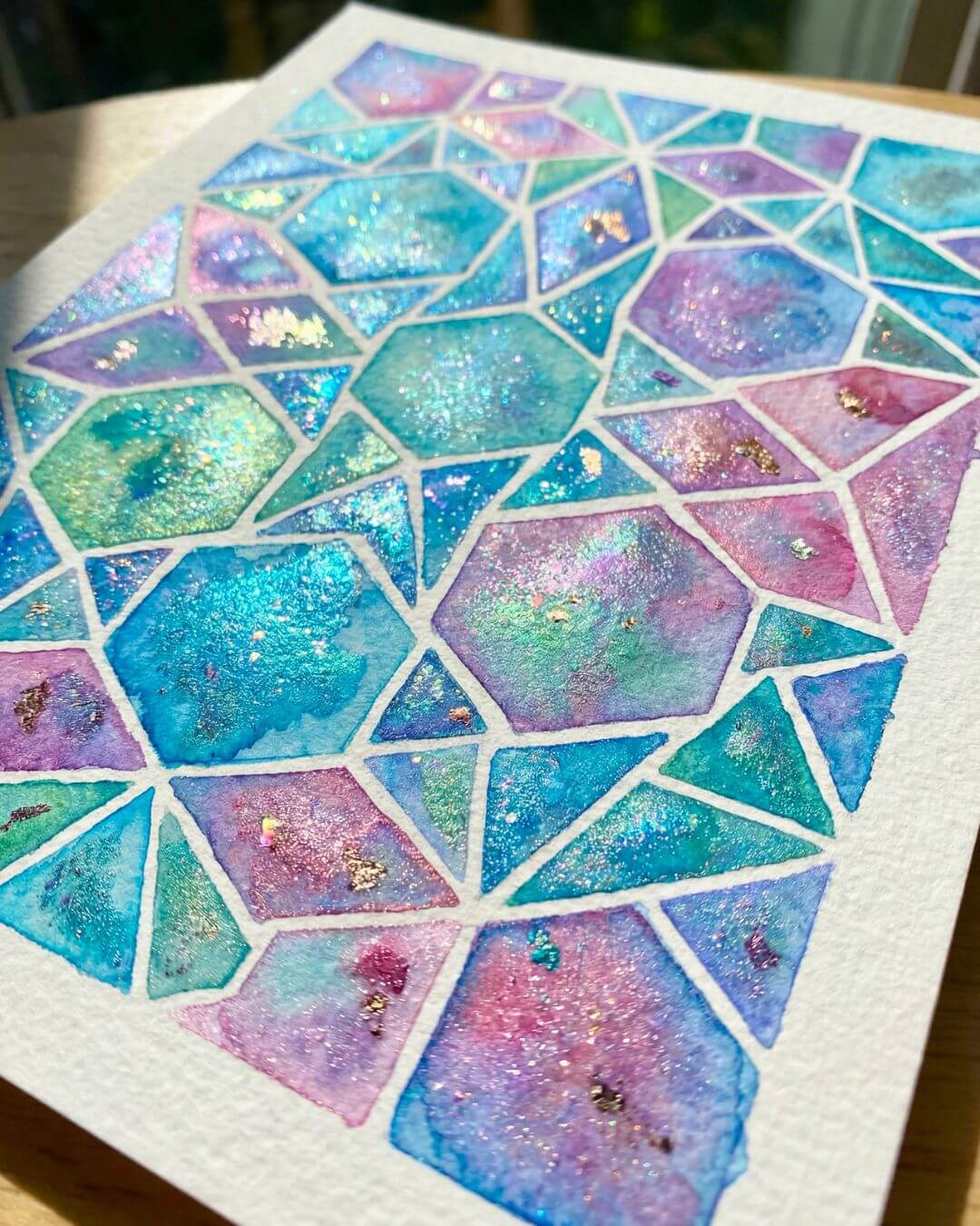 Close-up of a shimmering Islamic geometric watercolor painting in turquoise, blue, and soft pink tones with metallic sparkle on textured paper.