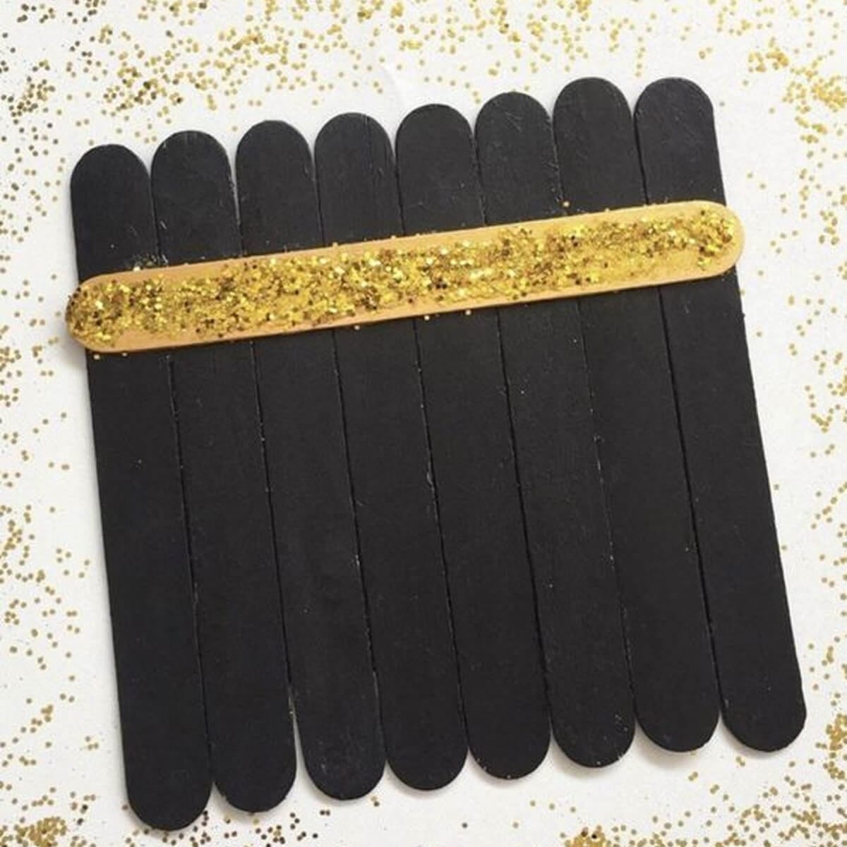 Easy Kaaba craft made from black-painted ice cream sticks with a gold glitter band on white paper.