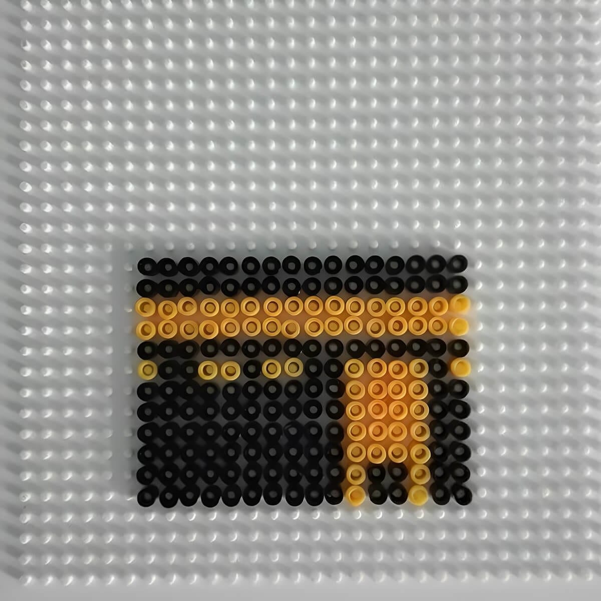Simple Kaaba design made with black and gold Perler beads on a white pegboard for a kids Islamic craft activity.