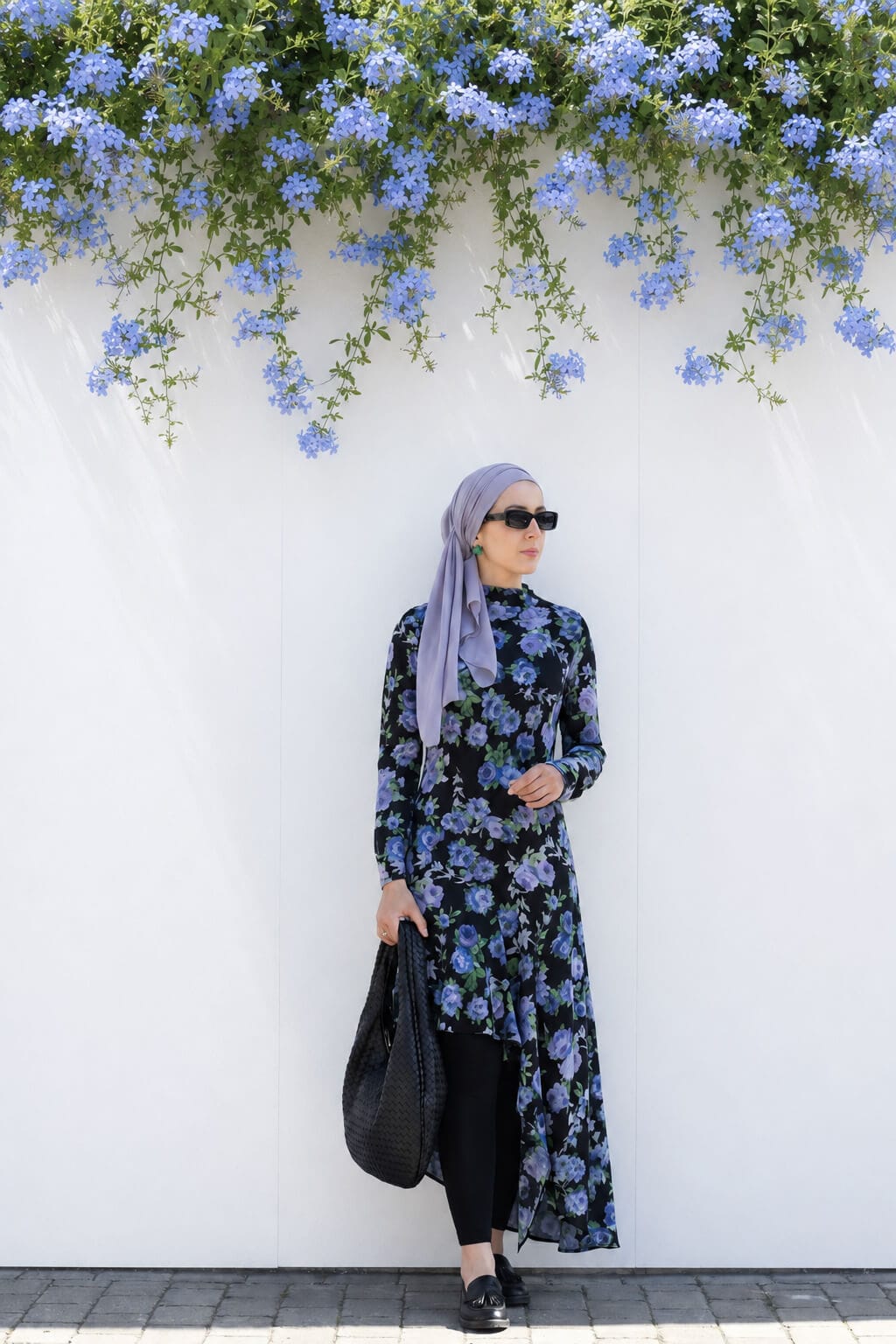 Hijabi woman standing by a white wall in a black floral asymmetric dress over leggings, styled with a lilac hijab, black loafers and a black woven shoulder bag.