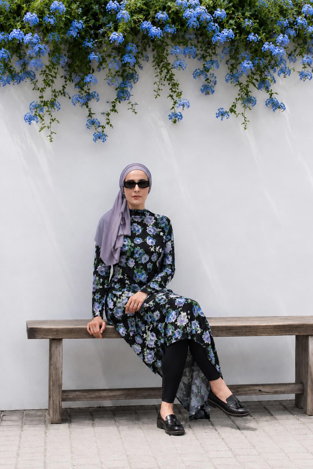 Hijabi woman sitting on a wooden bench wearing a black floral asymmetric dress over leggings with a lilac hijab.