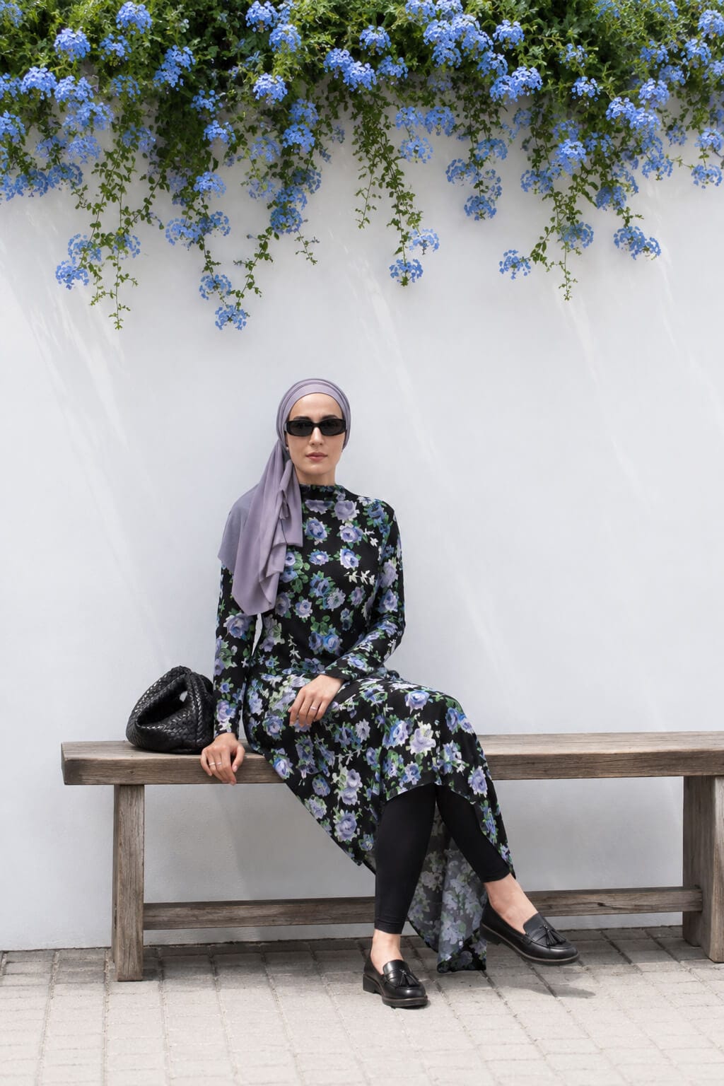 Hijabi woman sitting on a wooden bench wearing a black floral asymmetric dress over leggings with a lilac hijab, black loafers and shoulder bag.