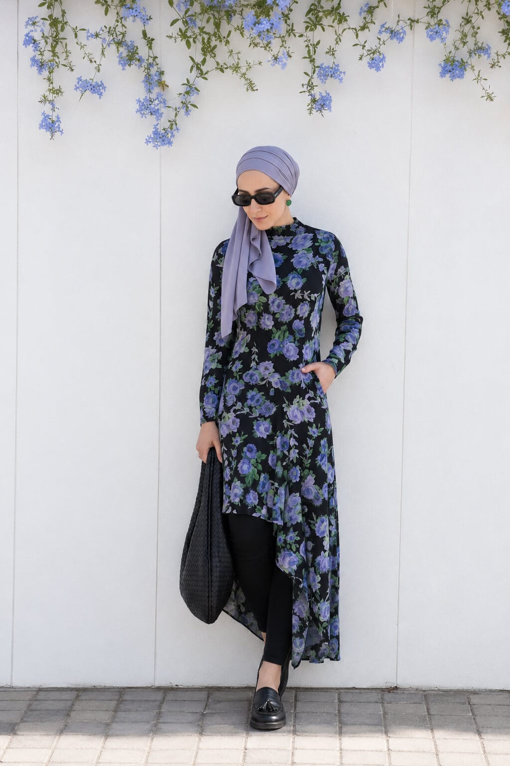 Hijabi woman standing by a white wall wearing a black floral asymmetric dress over leggings with a lilac hijab, black loafers and a black shoulder bag.