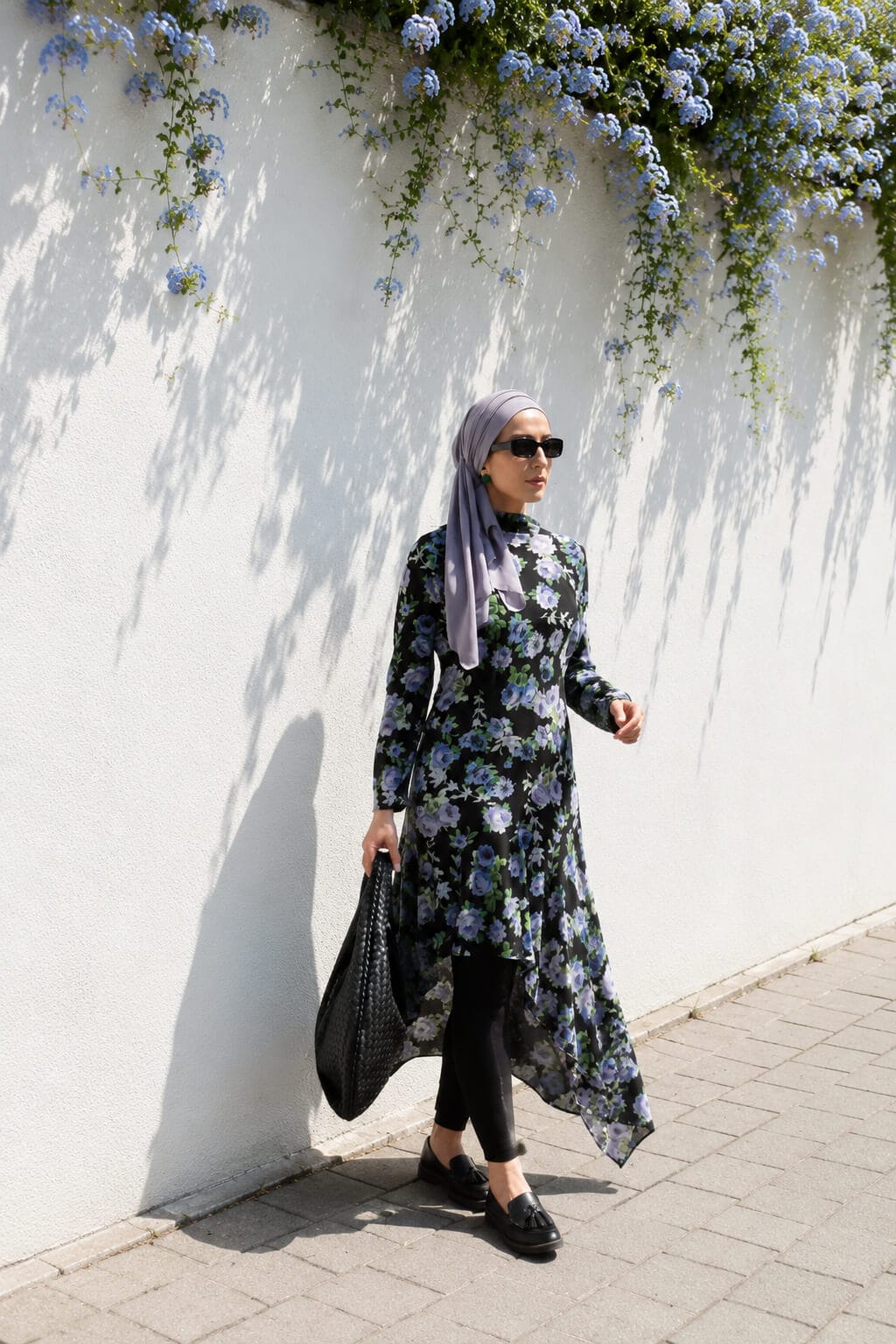 Hijabi woman walking by a white wall in a black floral asymmetric dress over leggings, styled with a lilac hijab, black loafers, sunglasses and a black shoulder bag.