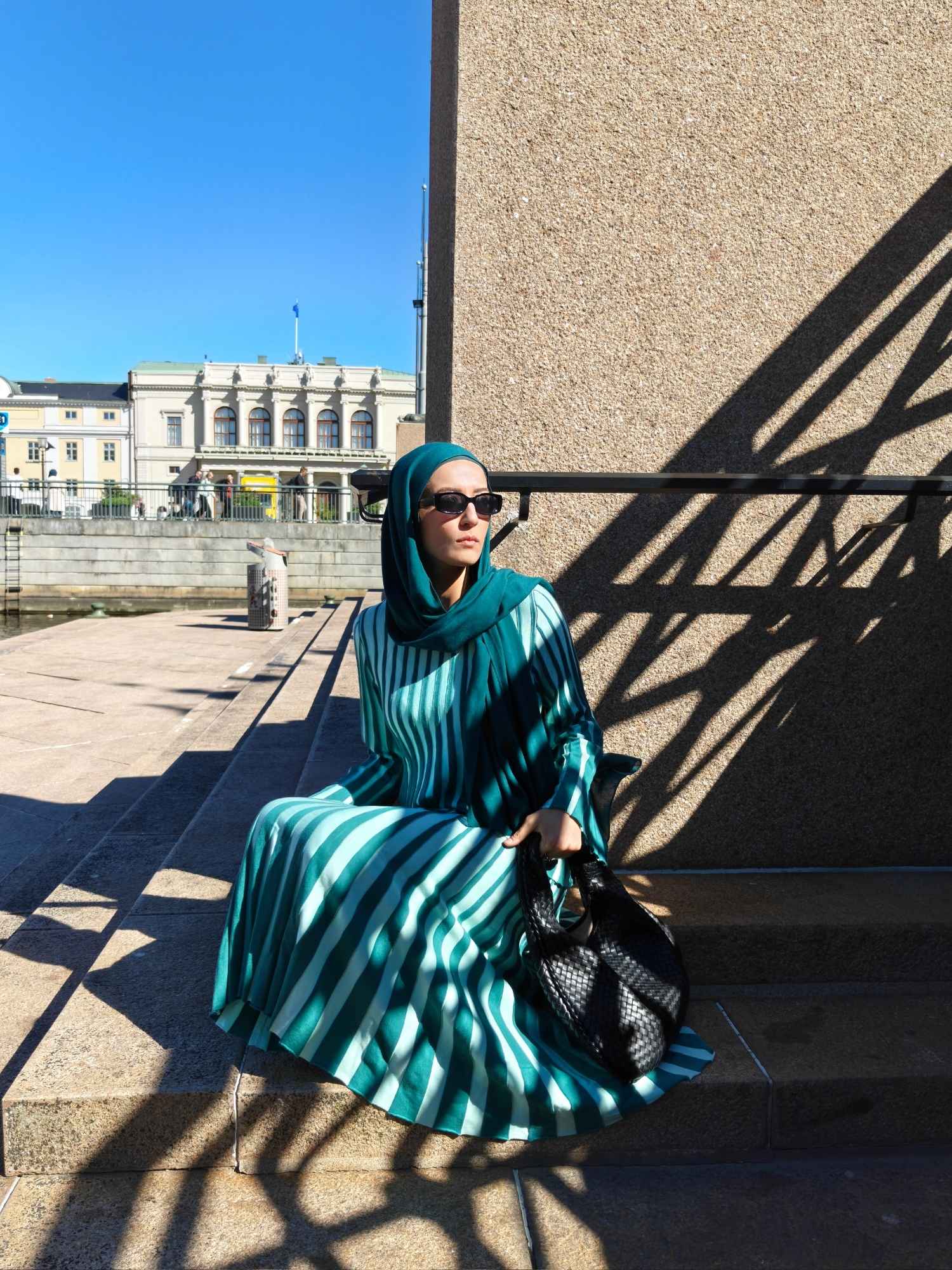 Hijabi woman sitting on city steps wearing a green pleated two-piece skirt set with matching hijab and woven black bag