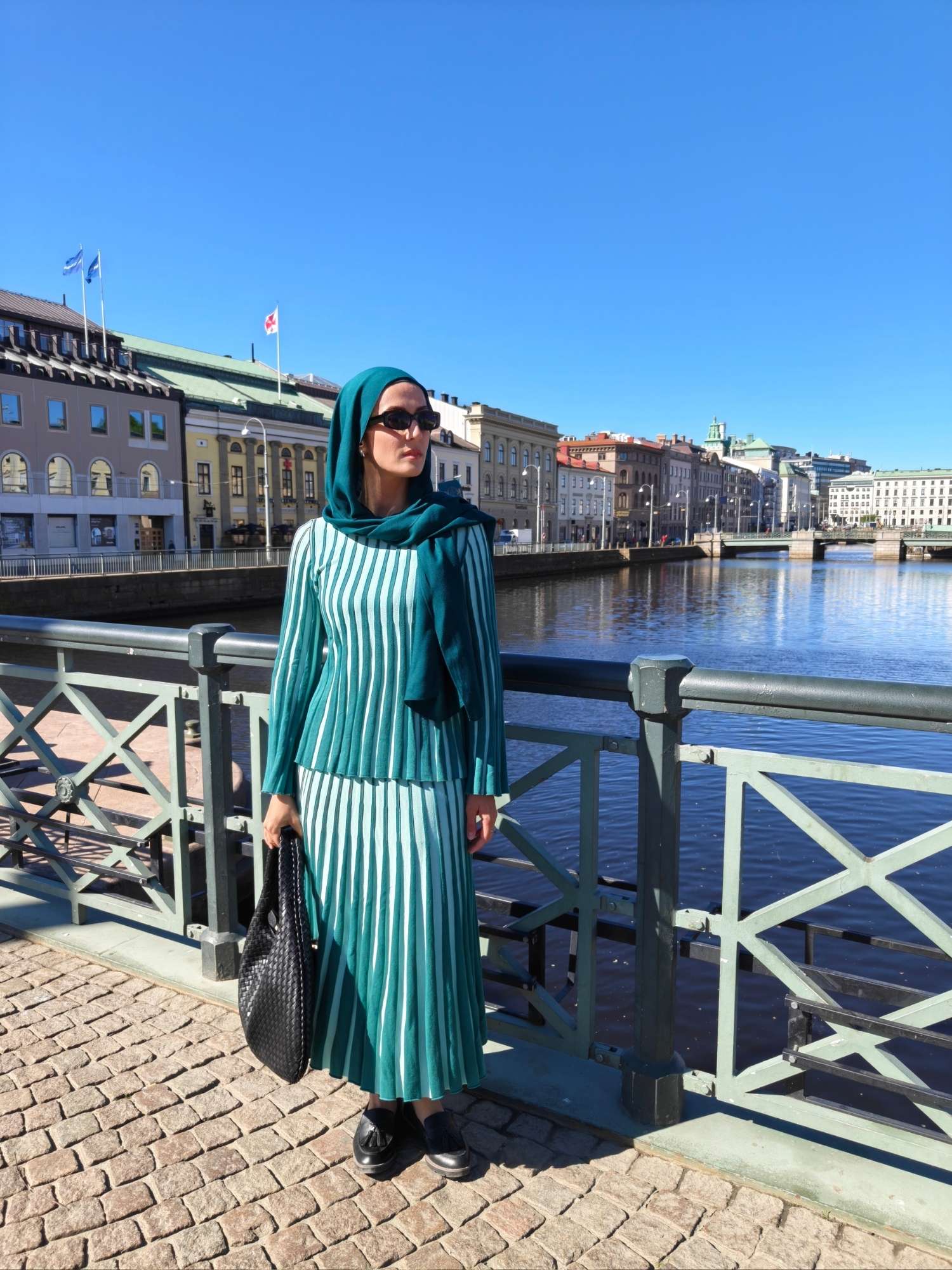 Hijabi woman wearing a green pleated two-piece skirt set with matching hijab, black loafers and woven bag by the canal in Gothenburg.