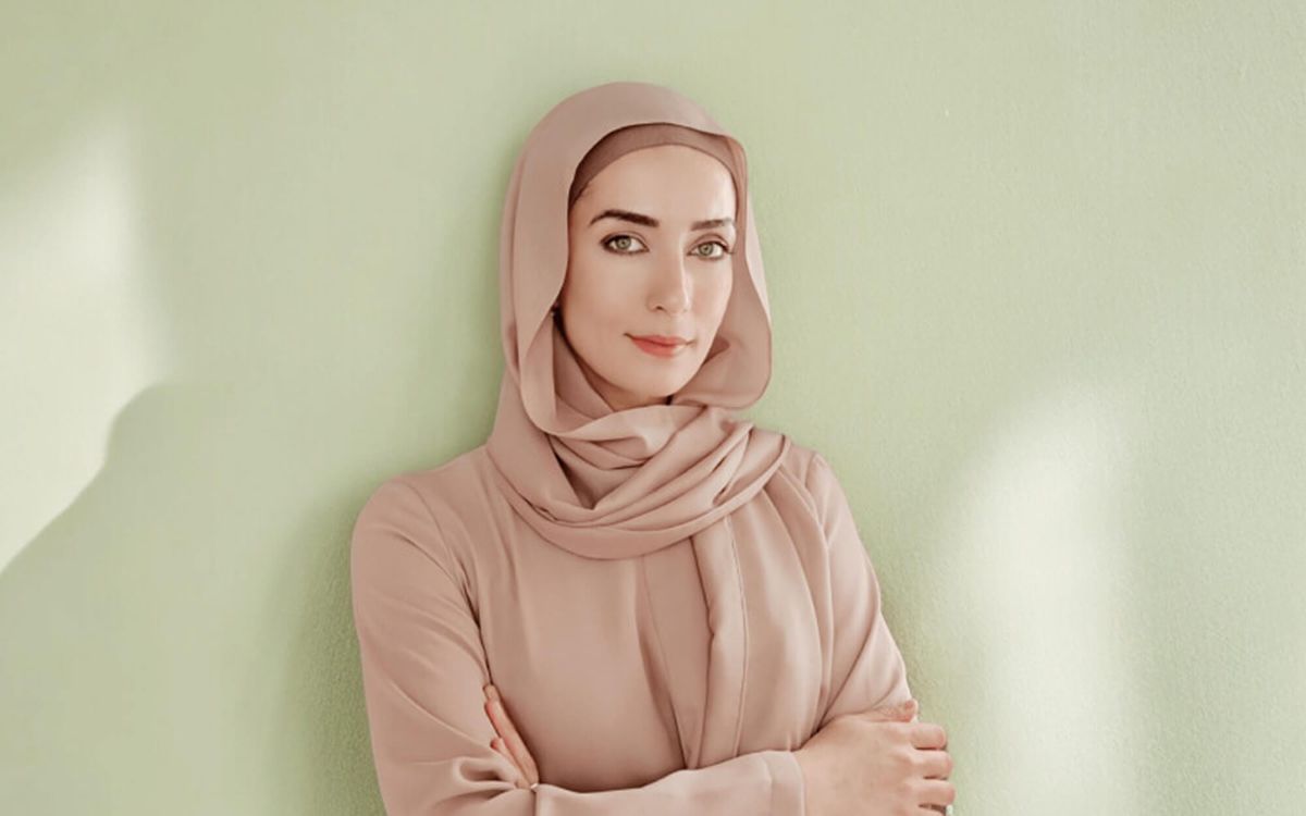 Portrait of Neda A. wearing a dusty rose hijab against a soft pastel green background.