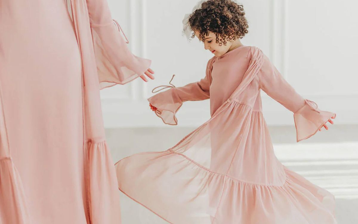 Mother and young daughter in matching soft pink, flowing modest dresses, the child gently twirling while holding the fabric of her dress in a bright, minimal room.