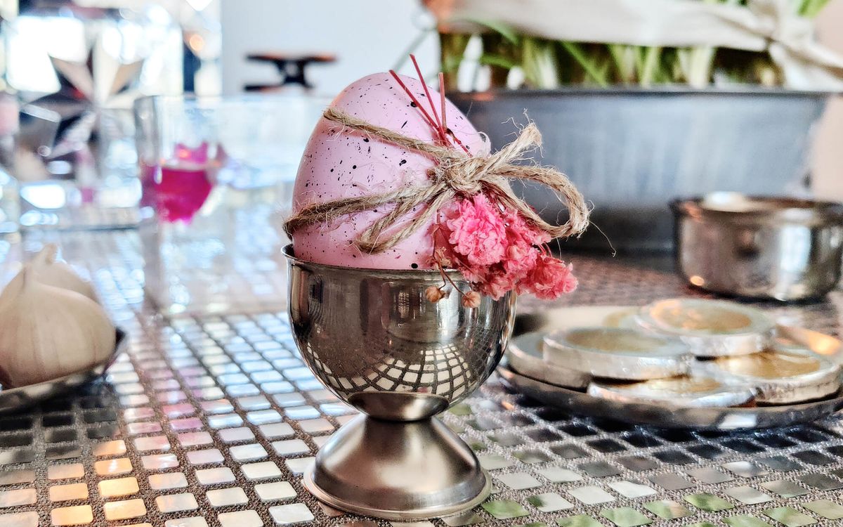 A softly speckled pink egg decorated with twine and small dried flowers, resting in a metal cup as part of a Haftseen display.