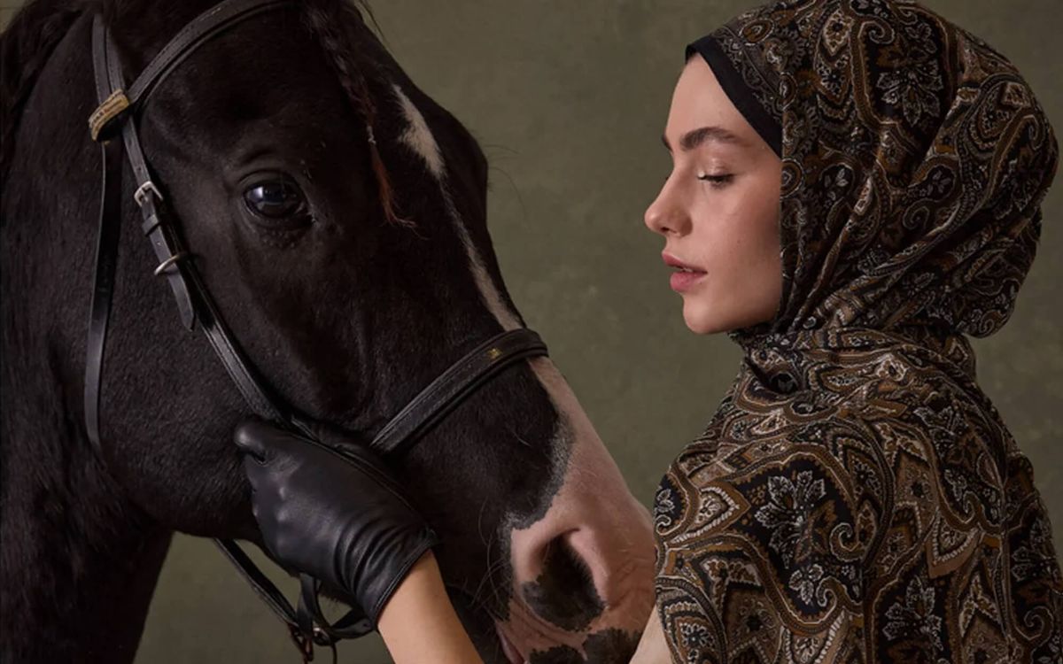 Woman in a paisley hijab gently holding a black horse’s face.