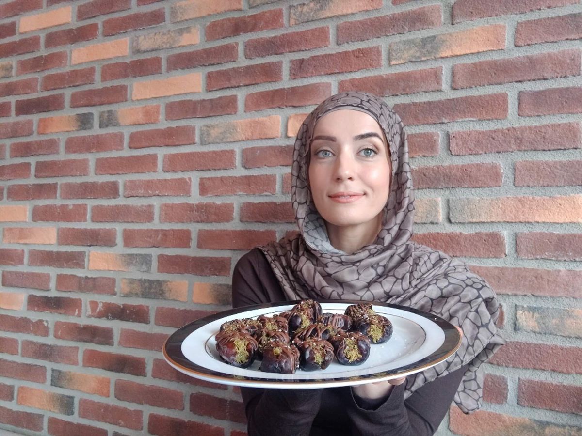 Woman in a patterned hijab holding a platter of Dubai-style chocolate-stuffed dates with Kataifi against a brick wall.