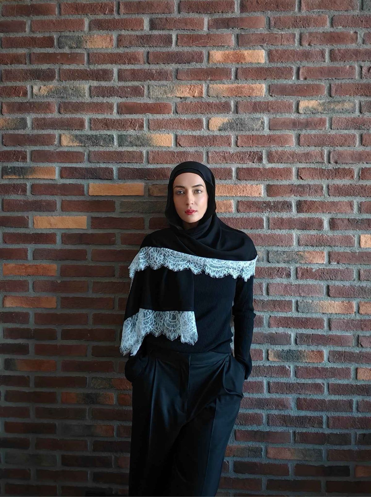 Woman wearing a black modal lace hijab styled with an all-black modest outfit against a brick wall