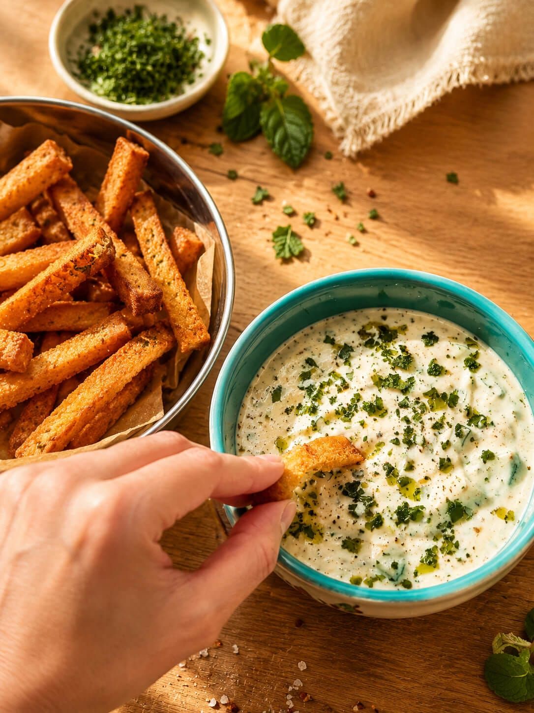 Hand dipping crispy leftover bread bite into mast o khiar yogurt dip topped with herbs and olive oil.
