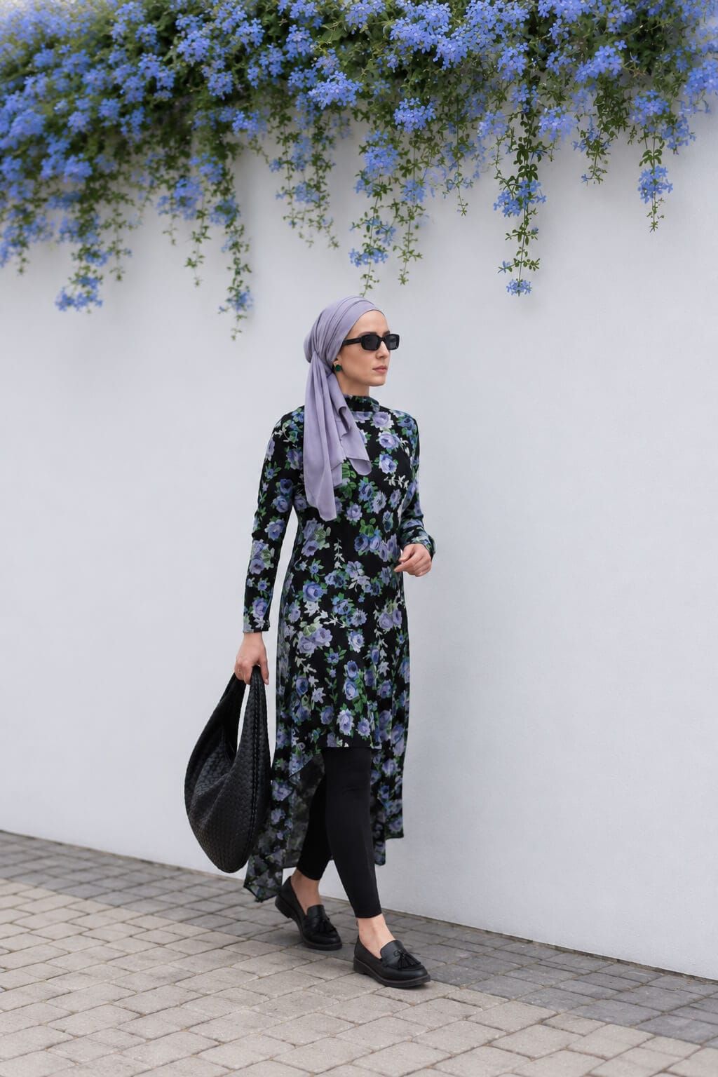 Hijabi woman wearing a black floral asymmetric dress over leggings with a lilac hijab, black loafers, sunglasses and a black shoulder bag as a modest summer outfit.
