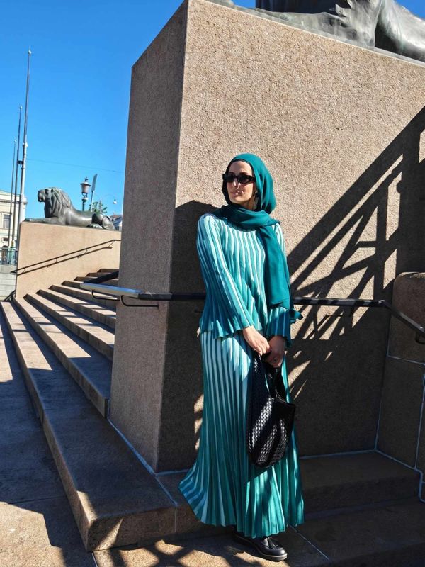 Hijabi woman wearing a green pleated two-piece skirt set with matching hijab, black loafers and woven bag in the city
