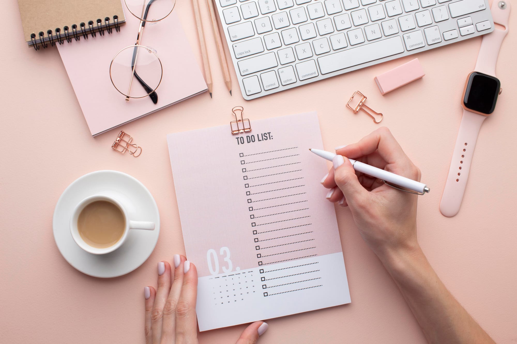 Vista superior de uma mão escrevendo em um papel de check list. Também há teclado, relógio, óculos e café sobre a mesa. 
