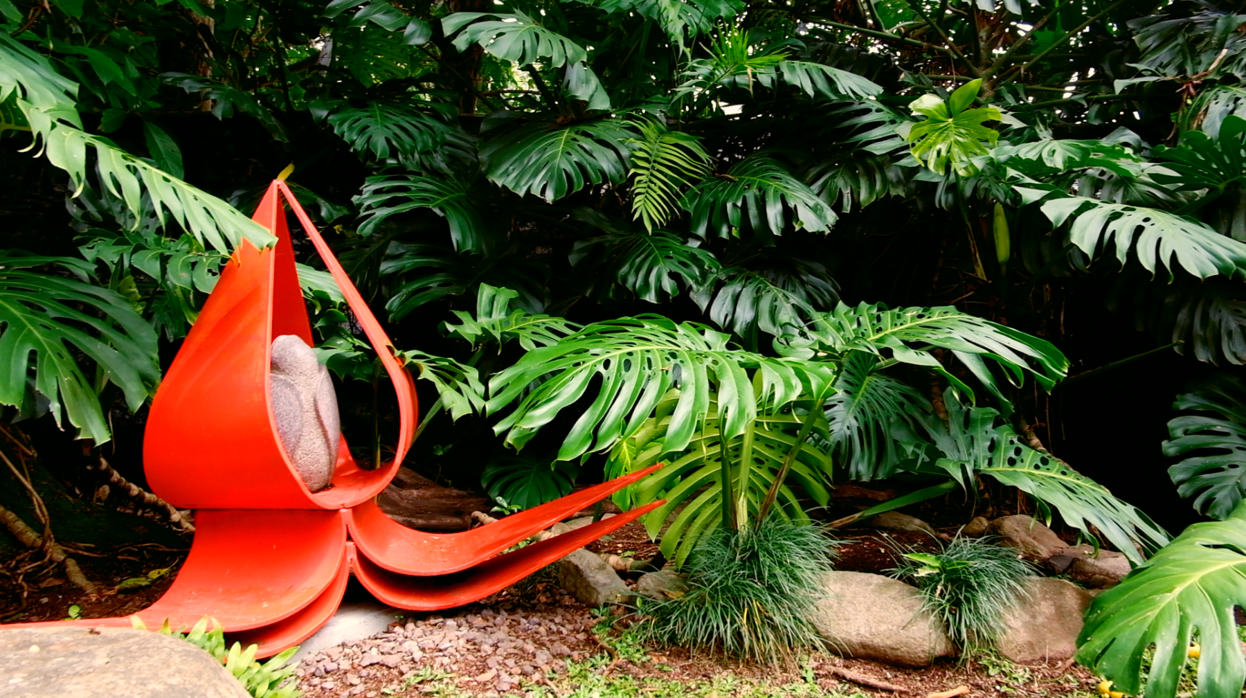 Una escultura de la flor de loto en rojo chroma que resalta por su tono saturado contra un fondo de naturaleza tropical verde. Escultor: José Sancho Benito