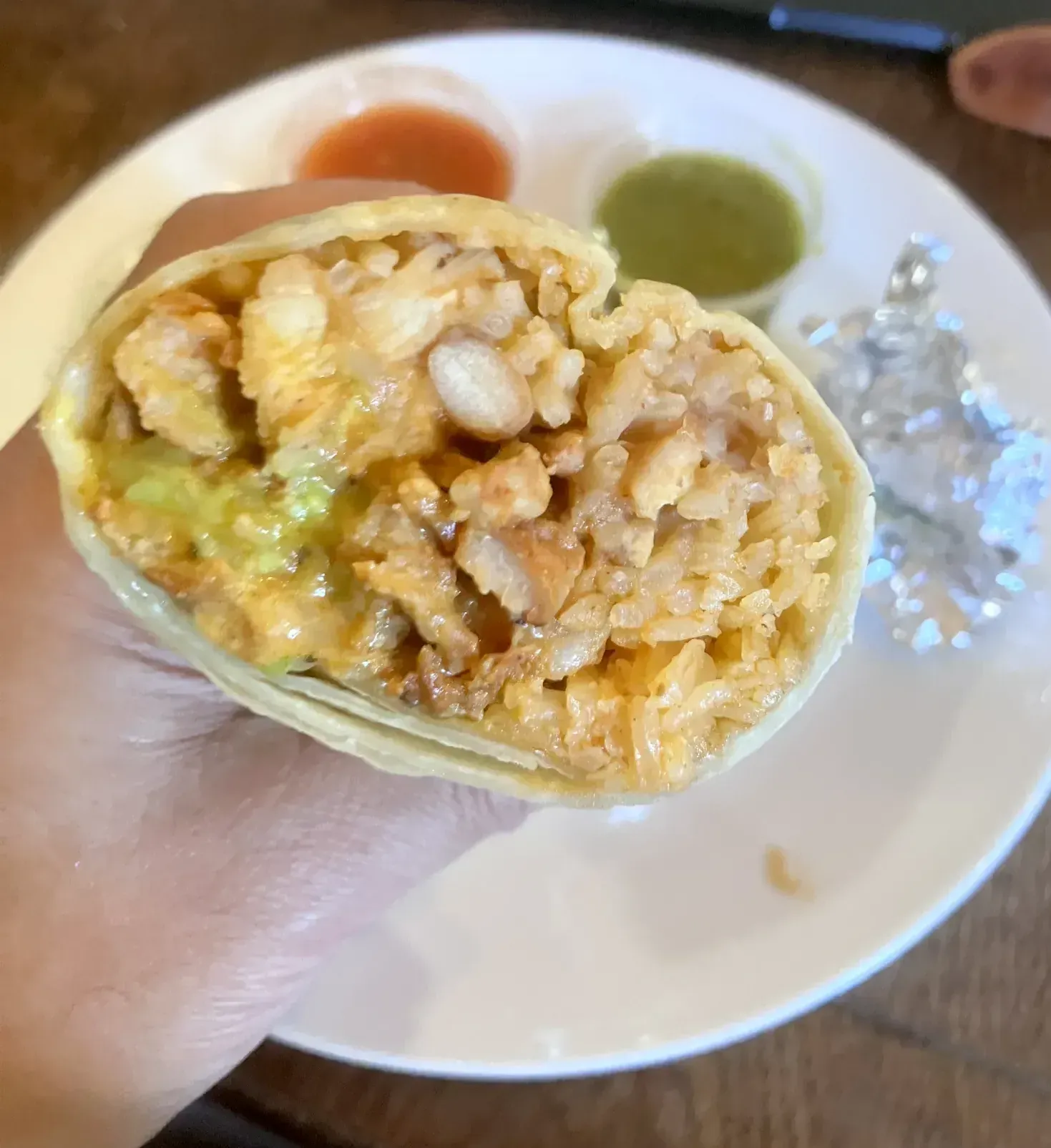 Hand holding a halved chicken burrito filled with rice, beans and chicken, with salsa cups on a plate behind it