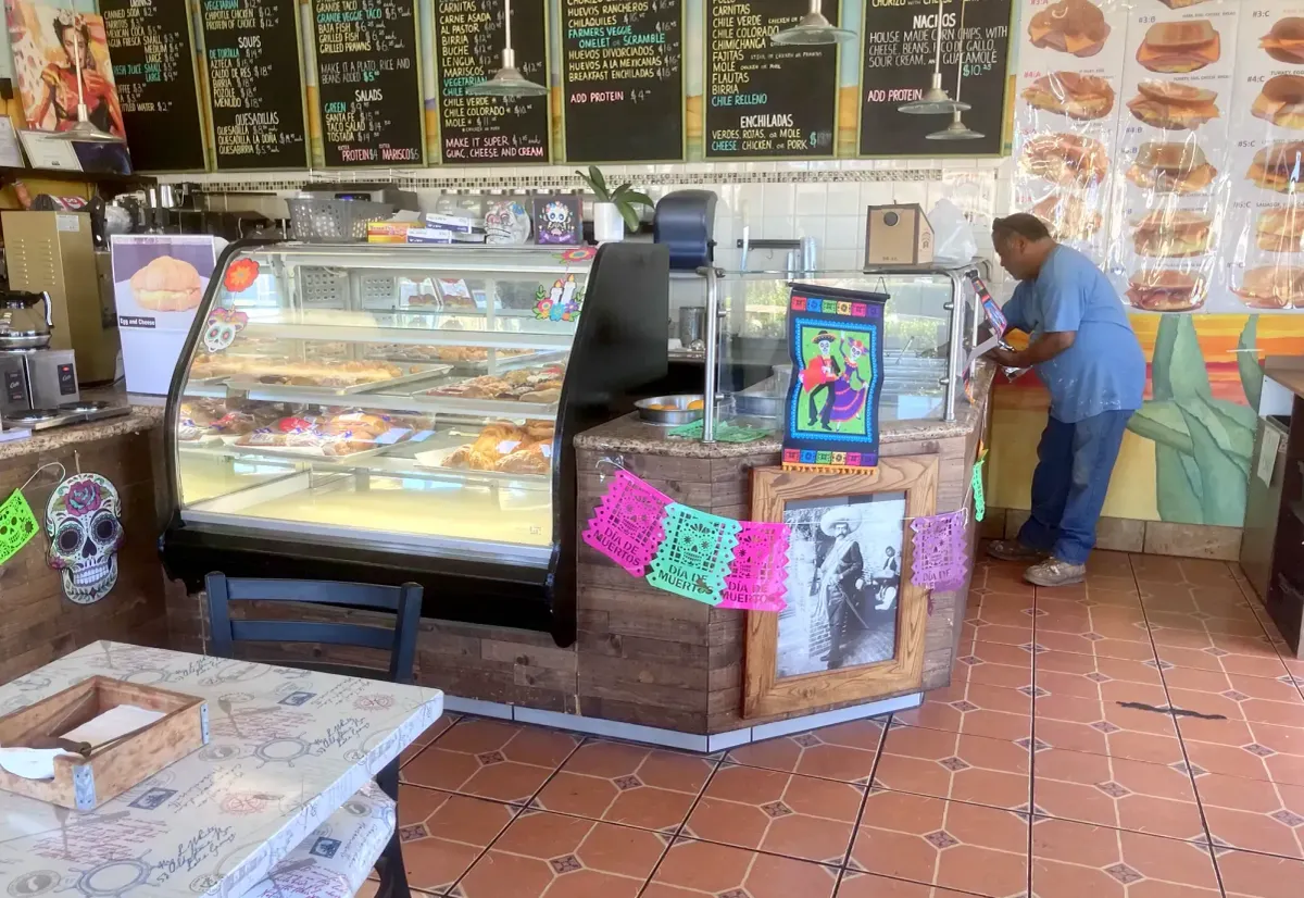 Man in a blue shirt at a taqueria counter beneath chalkboard menus, next to a pastry case and Día de Muertos decorations