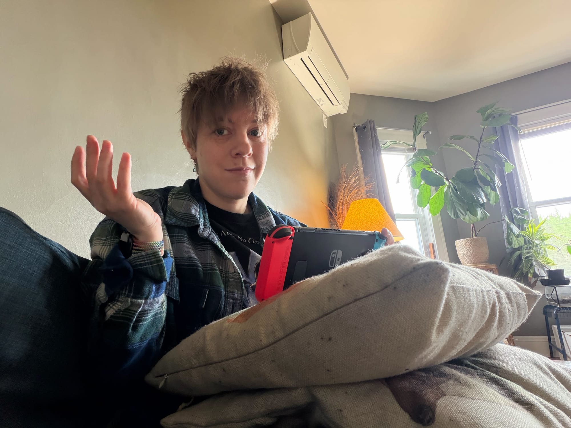 Maddy sits with two regular pillows stacked on one another and the Switch resting on top. She holds one hand up.