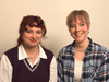 Zoë sits to the left in a collared shirt, smiling. Maddy sits to the right in a flannel shirt, smiling.