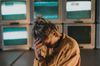 A person with their head in their hand, looking downcast, in front of a wall of TVs