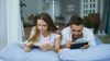 A woman and man lying on a bed together. The woman is reading a book and the man is tapping something on a tablet.